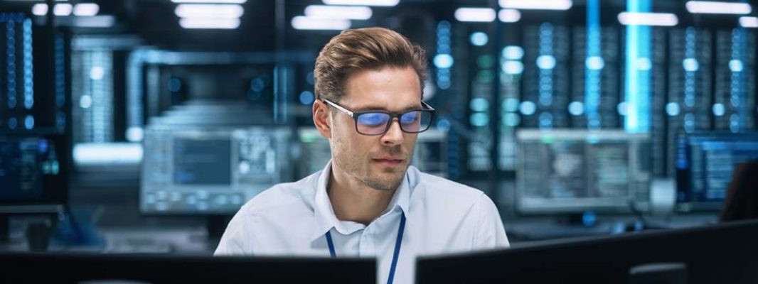 Administrator_Leadimage Ein Mann mit Brille sitzt vor Bildschirmen mit Servern im Hintergrund.